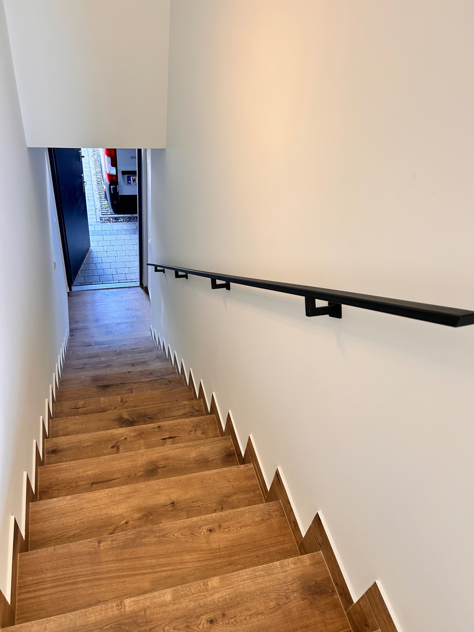 Drewniane schody z czarną, minimalistyczną poręczą zamontowaną na białej ścianie. Widok z góry na dół, w tle otwarte drzwi i fragment zaparkowanego samochodu.