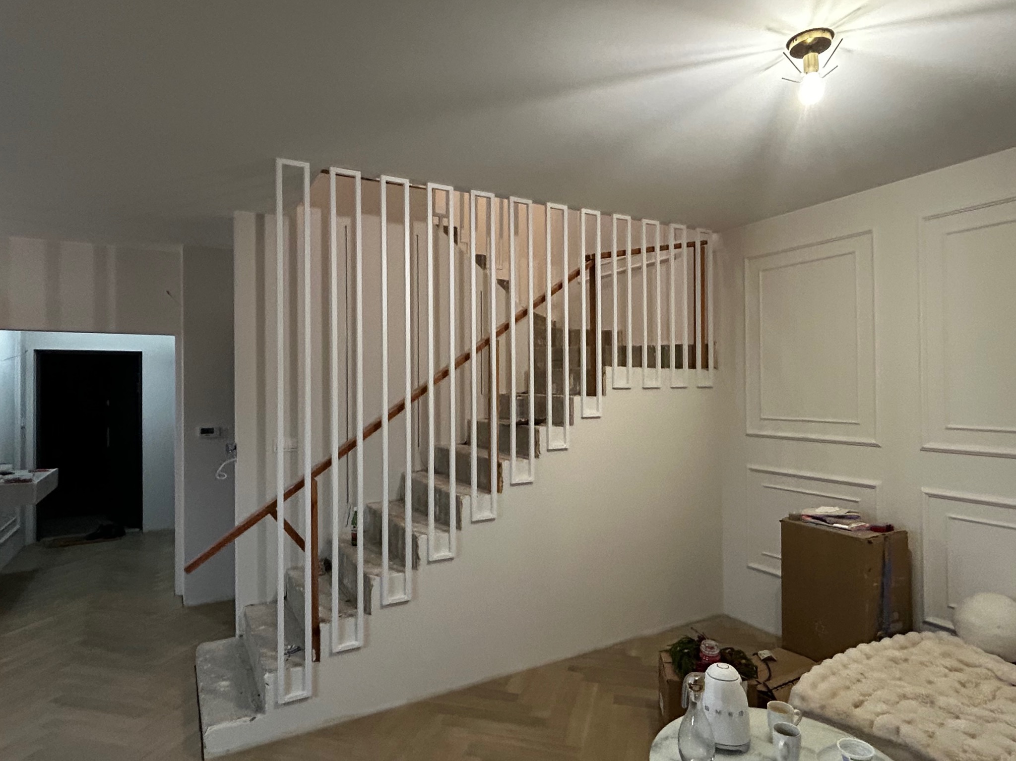 Nowoczesne schody wewnętrzne z białą, minimalistyczną balustradą z pionowych elementów i drewnianą poręczą. Widok na jasne wnętrze domu w trakcie wykończenia.