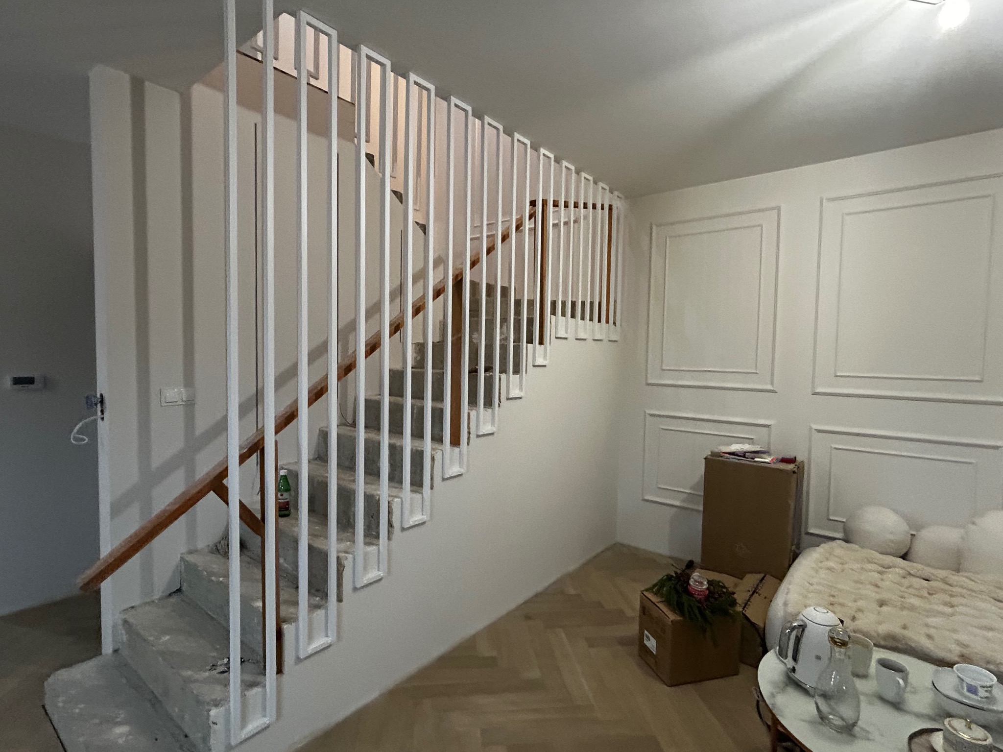 Nowoczesne schody wewnętrzne z białą, minimalistyczną balustradą metalową i drewnianym poręczą. Widok na jasne wnętrze z parkietem i dekoracyjną ścianą.