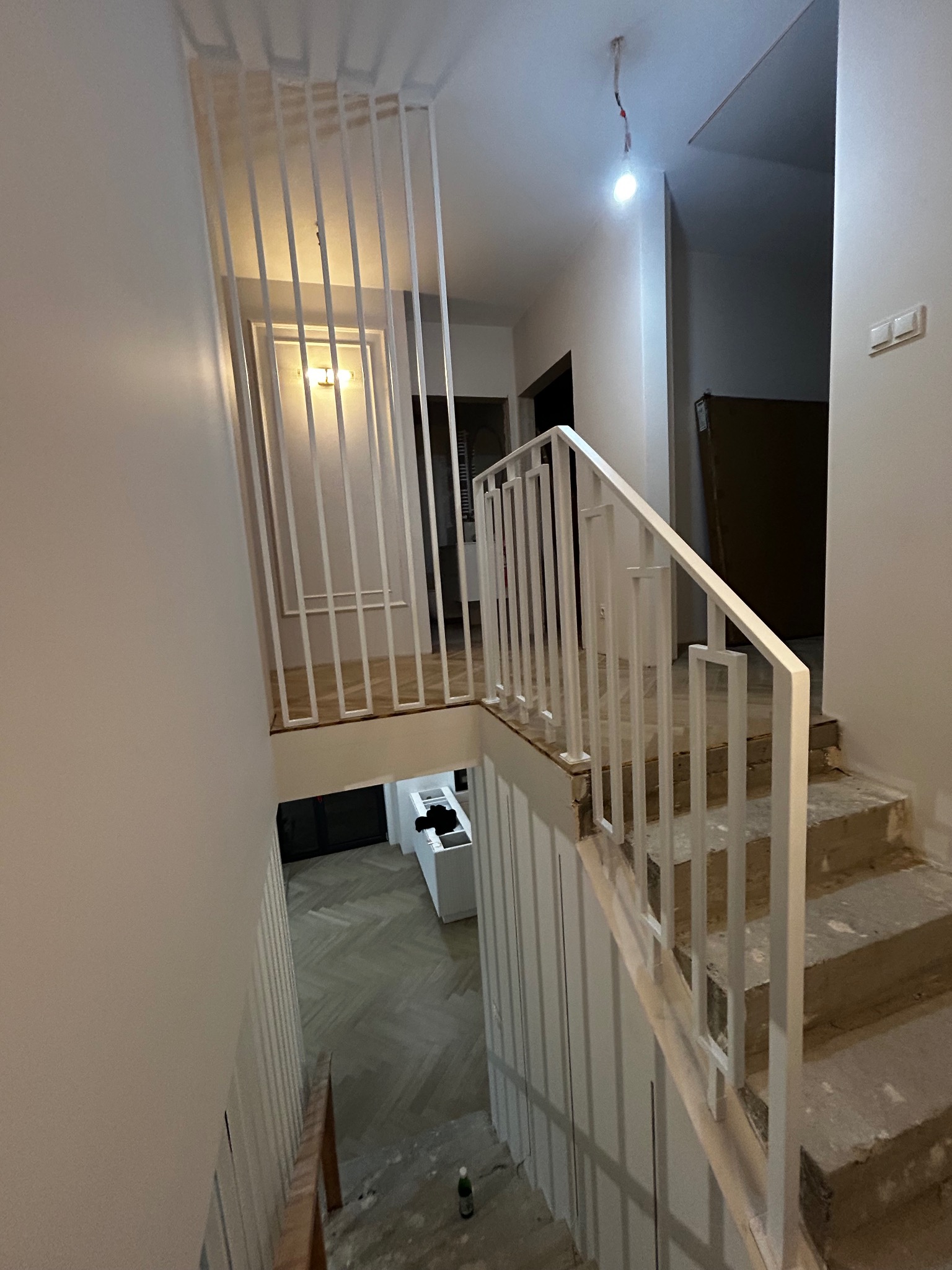 Biała, nowoczesna balustrada schodowa z pionowymi elementami, widok z góry na schody w trakcie remontu. Minimalistyczny design, surowe wykończenie stopni.