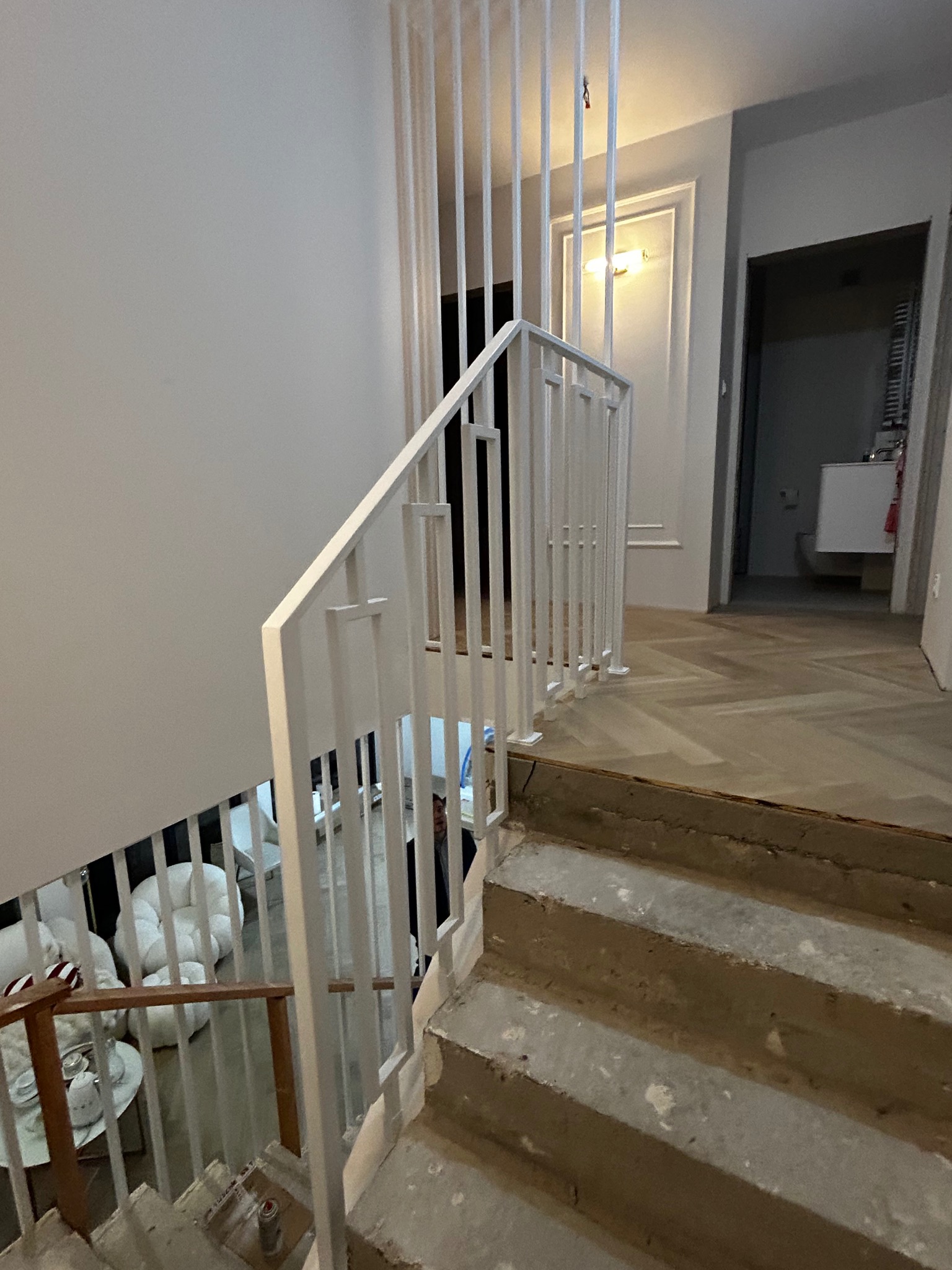 Biała balustrada schodowa z pionowymi szczeblami w nowoczesnym wnętrzu. Widok z góry na schody w trakcie wykończenia, z widocznym fragmentem salonu i łazienki w tle.