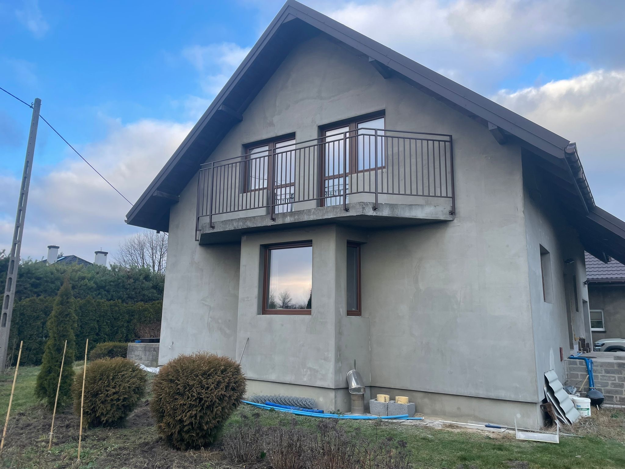 Szary dom jednorodzinny z balkonem i brązową, metalową balustradą. Widoczne okna z brązowymi ramami i fragment ogrodu z roślinami.