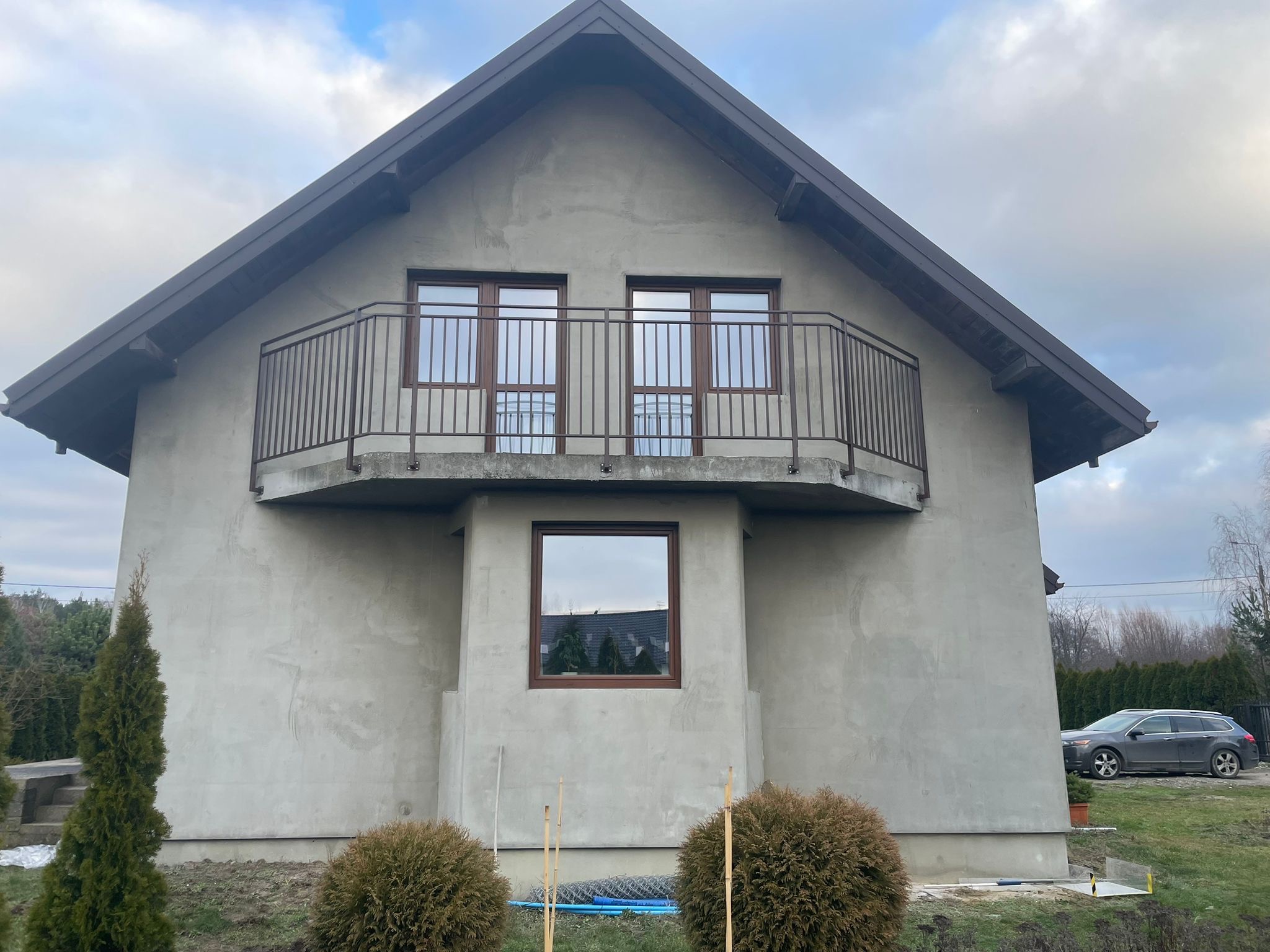 Surowy dom z brązową, metalową balustradą balkonową na pierwszym piętrze. Widoczne okna z brązowymi ramami i fragment ogrodu z krzewami. Dom w trakcie wykończenia.