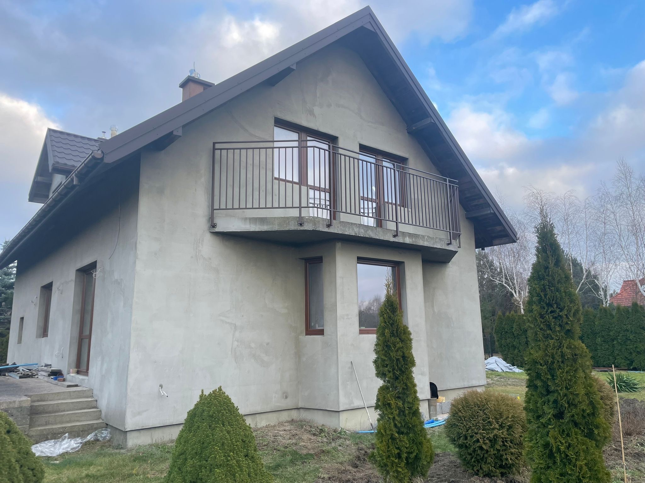 Dom w stanie surowym z brązową, metalową balustradą balkonową. Widoczne okna z brązowymi ramami i fragment schodów wejściowych. Tło stanowi pochmurne niebo.