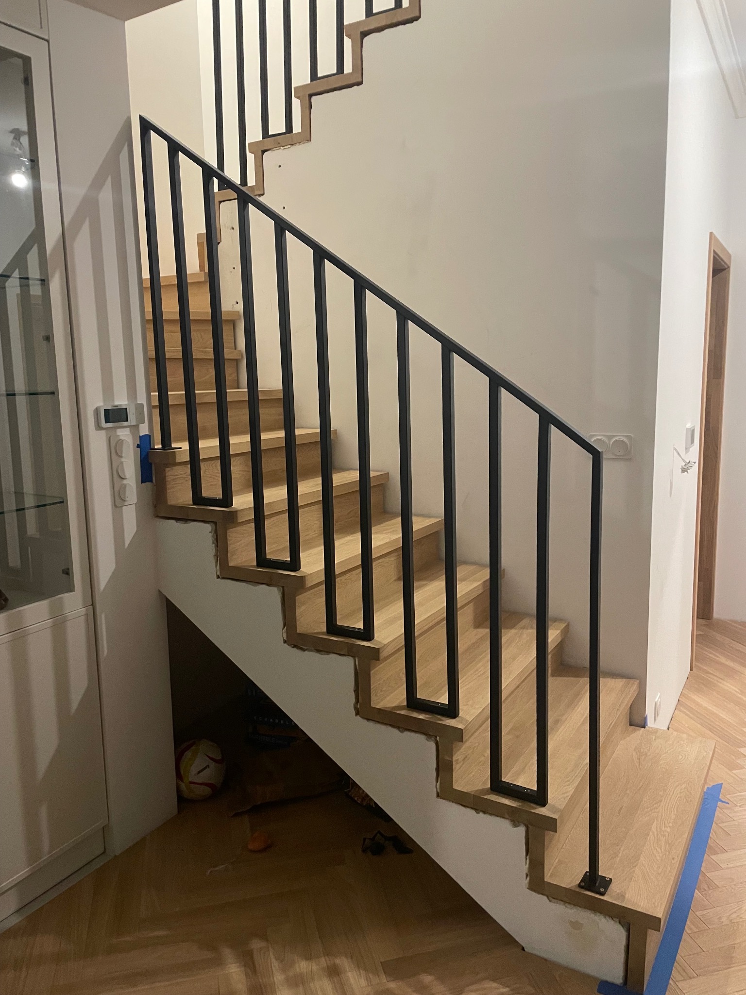 Drewniane schody z czarną, minimalistyczną balustradą metalową. Widoczne detale wykończenia i parkiet w jodełkę. Pod schodami piłka.