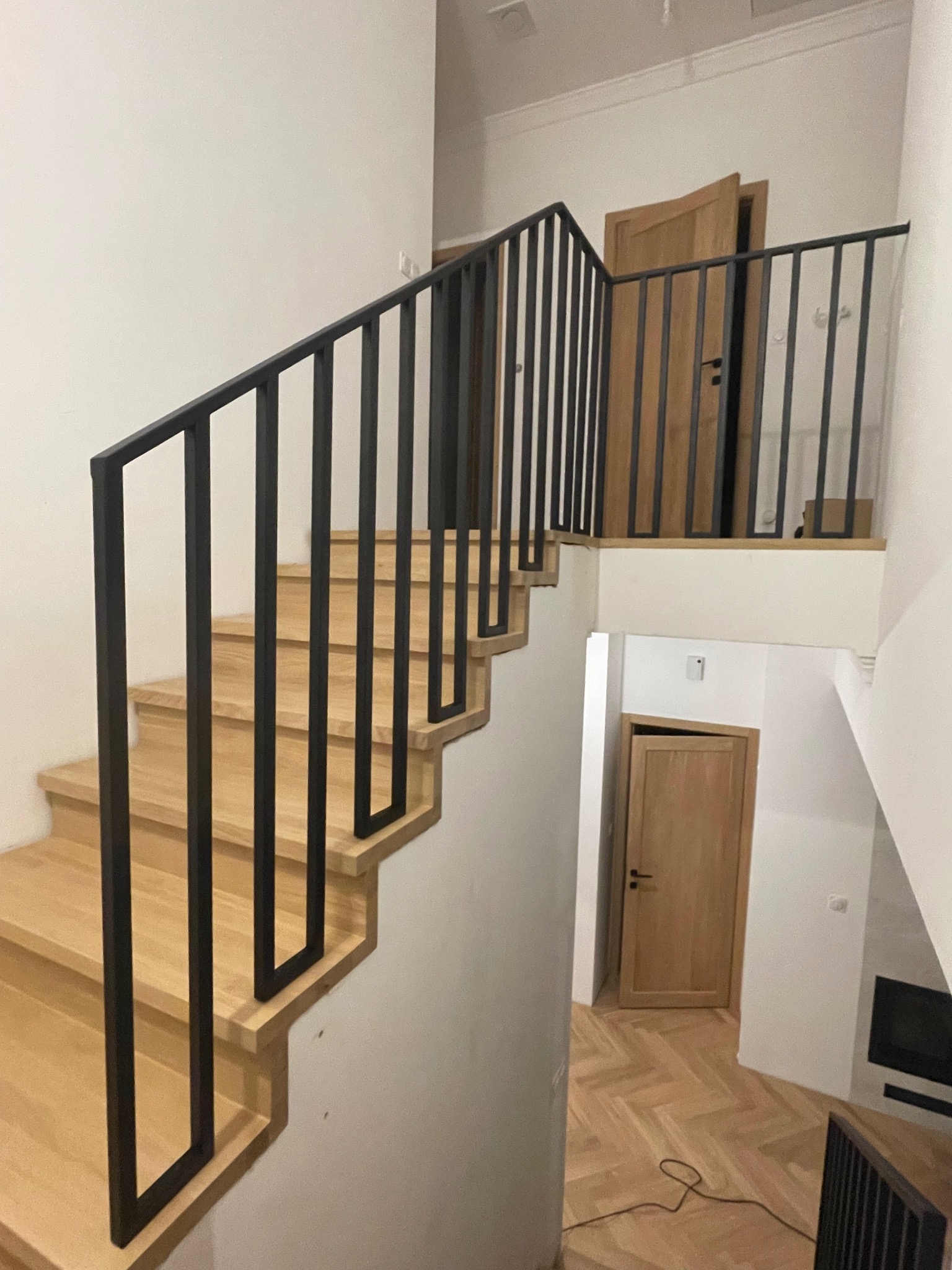 Nowoczesne schody drewniane z czarną, metalową balustradą o minimalistycznym designie. Widok z dołu, ukazujący konstrukcję i wykończenie. Jasne drewno kontrastuje z czernią.