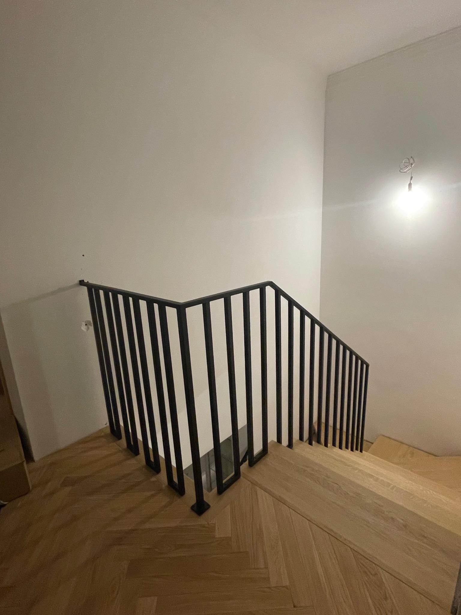 Minimalistyczna, czarna balustrada metalowa z pionowymi szczeblami, zamontowana na drewnianych schodach w jasnym wnętrzu. Ujęcie z góry, widoczny fragment parkietu.