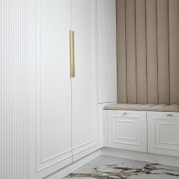 Meble A&K Arkadiusz Madaliński - Biała szafa wnękowa z pionowymi żłobieniami i złotymi uchwytami, uzupełniona siedziskiem z tapicerowanym panelem ściennym w beżowym odcieniu, na podłodze marmurowe płytki.