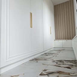 Meble A&K Arkadiusz Madaliński - Długi korytarz z zabudową szaf w kolorze białym, z frezowanymi frontami i podłużnymi złotymi uchwytami, marmurowa podłoga z brązowymi żyłami, na końcu siedzisko z tapicerowanym panelem ściennym...