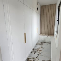 Meble A&K Arkadiusz Madaliński - Długa, biała szafa wnękowa z ozdobnymi frezami i złotymi uchwytami, dopasowana do wnęki z tapicerowanym siedziskiem i panelem ściennym, na podłodze marmurowe płytki.