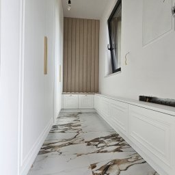 Meble A&K Arkadiusz Madaliński - Długi, wąski korytarz z białymi szafami wnękowymi po lewej stronie, siedziskiem przy oknie z tapicerowanym panelem na ścianie, białą podłogą z marmurkowym wzorem i niedokończonymi elementami...