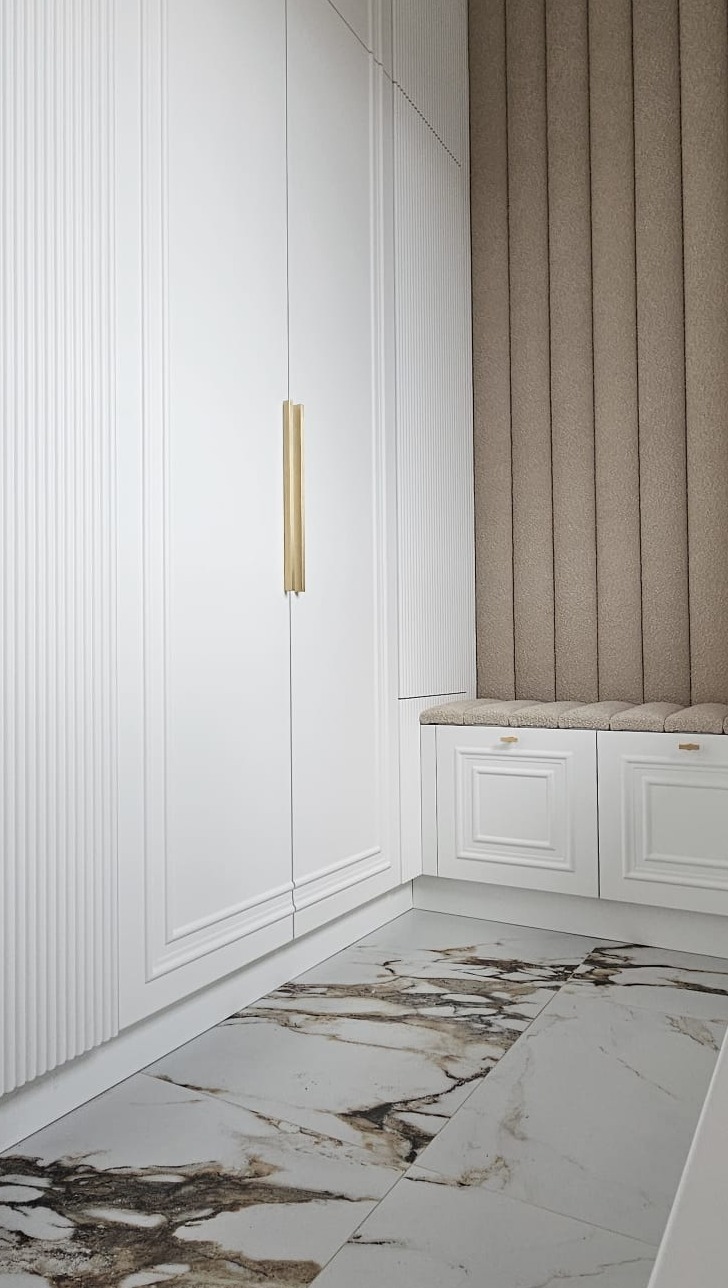 Biała szafa wnękowa z pionowymi żłobieniami i złotymi uchwytami, uzupełniona siedziskiem z tapicerowanym panelem ściennym w beżowym odcieniu, na podłodze marmurowe płytki.