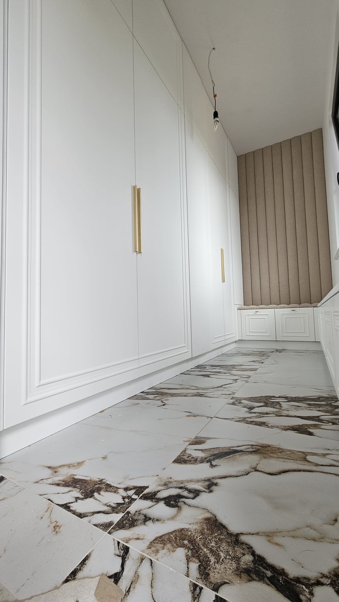 Długi korytarz z zabudową szaf w kolorze białym, z frezowanymi frontami i podłużnymi złotymi uchwytami, marmurowa podłoga z brązowymi żyłami, na końcu siedzisko z tapicerowanym panelem ściennym...