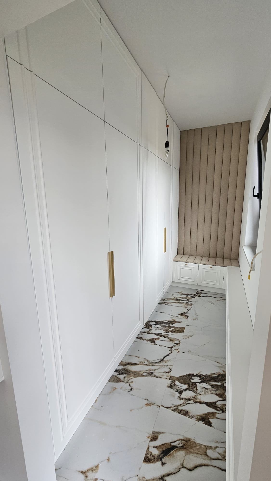 Długa, biała szafa wnękowa z ozdobnymi frezami i złotymi uchwytami, dopasowana do wnęki z tapicerowanym siedziskiem i panelem ściennym, na podłodze marmurowe płytki.