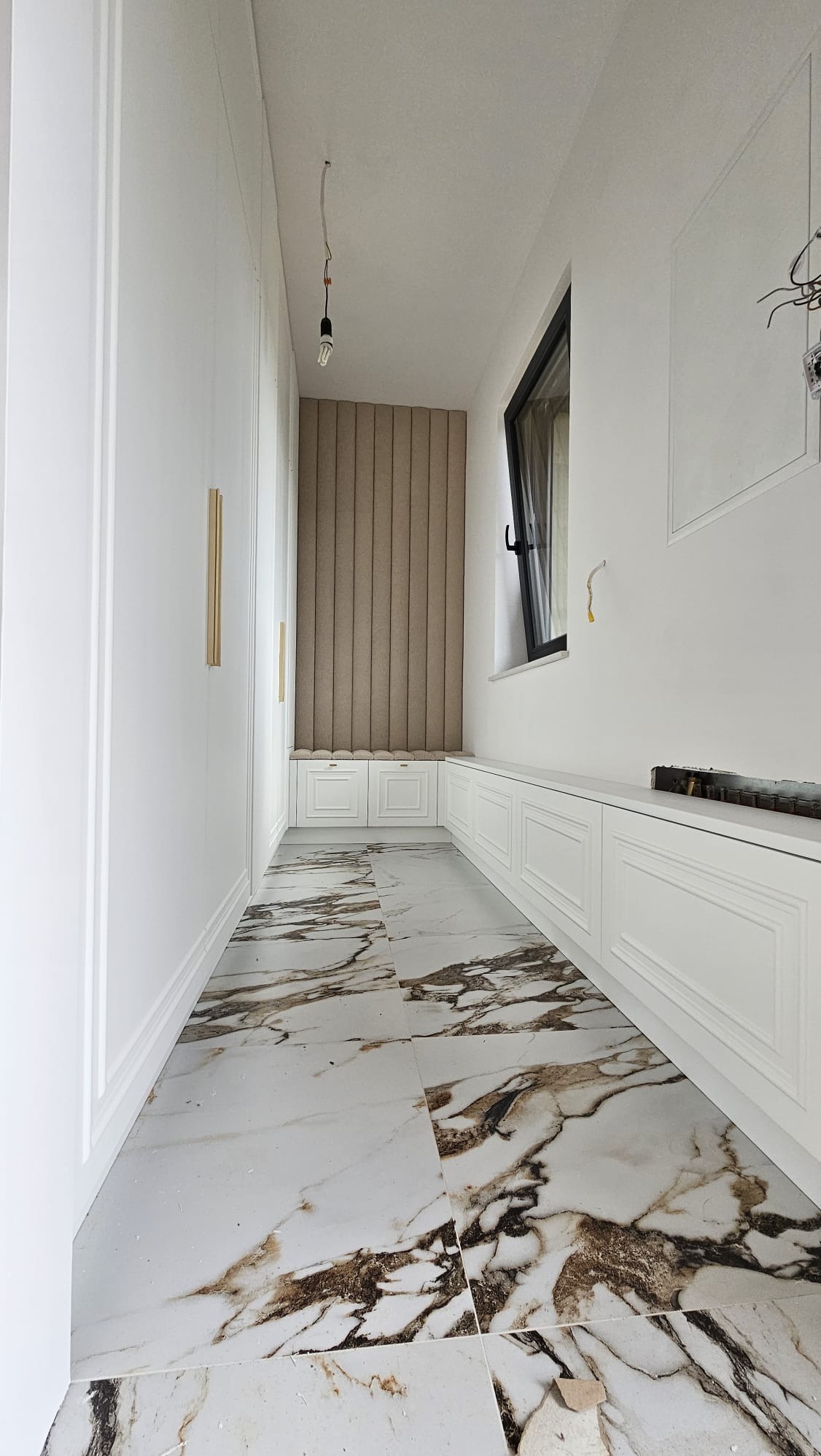 Długi, wąski korytarz z białymi szafami wnękowymi po lewej stronie, siedziskiem przy oknie z tapicerowanym panelem na ścianie, białą podłogą z marmurkowym wzorem i niedokończonymi elementami...
