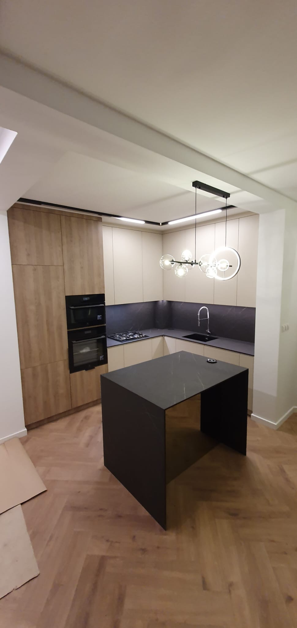 Nowoczesna kuchnia z wyspą, fronty szafek w odcieniach beżu i drewna, czarny blat i okładzina ściany, designerska lampa z kulistymi kloszami.