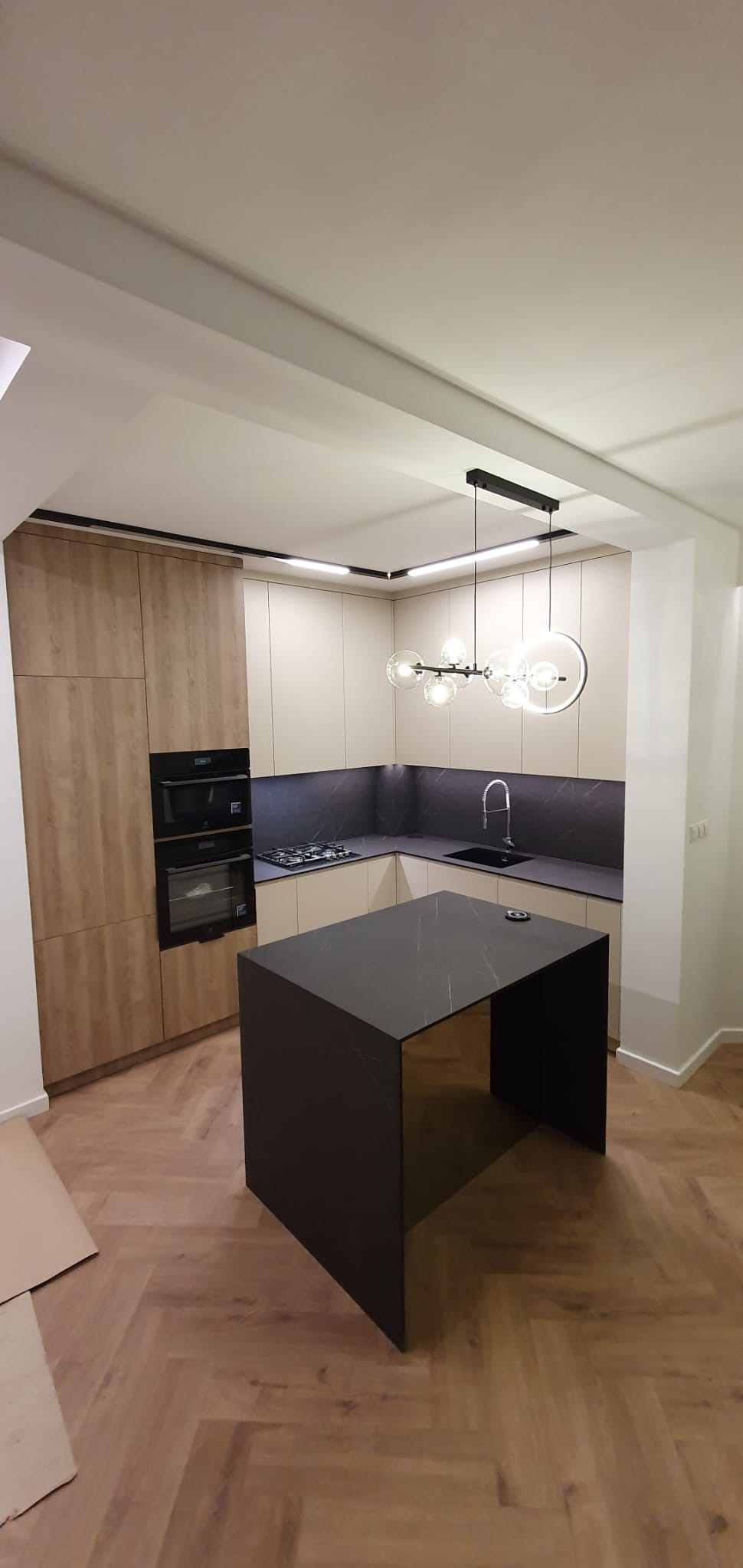 Nowoczesna kuchnia z wyspą o czarnym blacie, jasne szafki, drewniana podłoga w jodełkę, oświetlenie punktowe i designerska lampa wisząca.