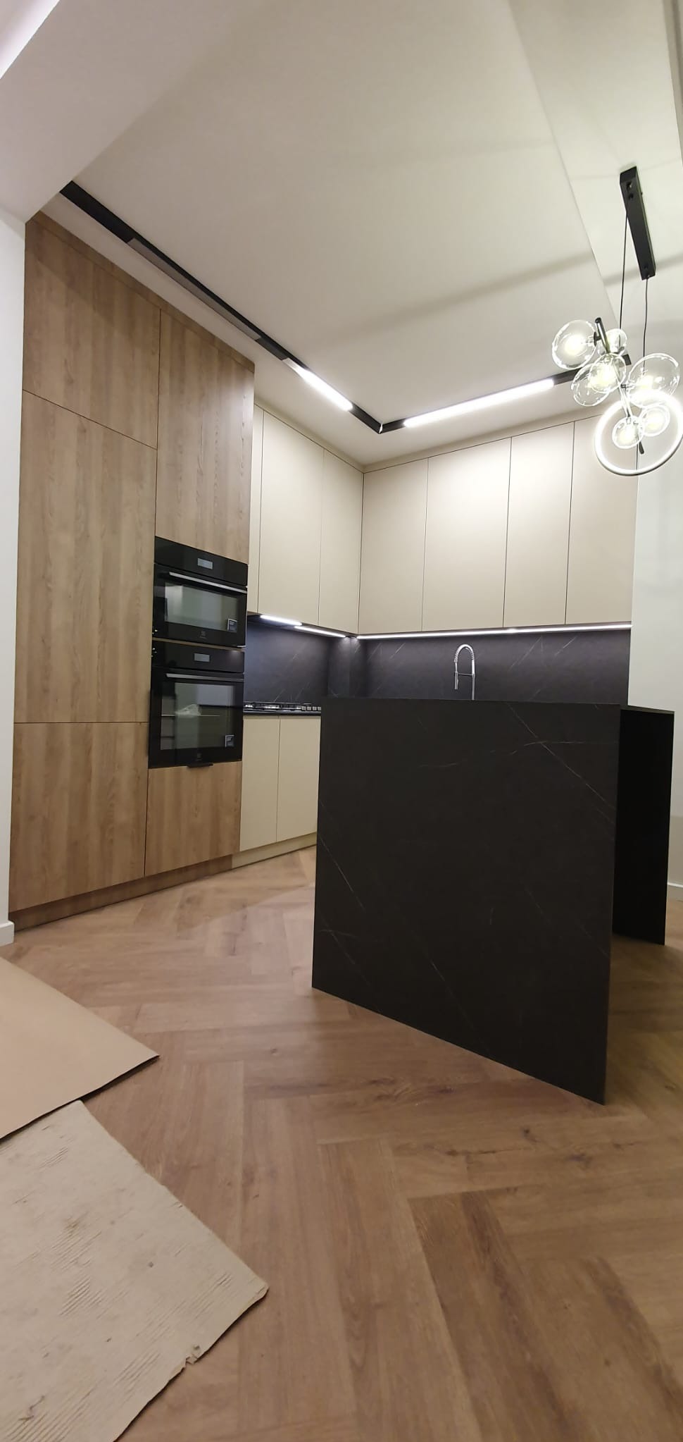 Nowoczesna kuchnia z wyspą i zabudową drewnopodobną, oświetlenie podszafkowe, parkiet w jodełkę, kartony ochronne na podłodze.