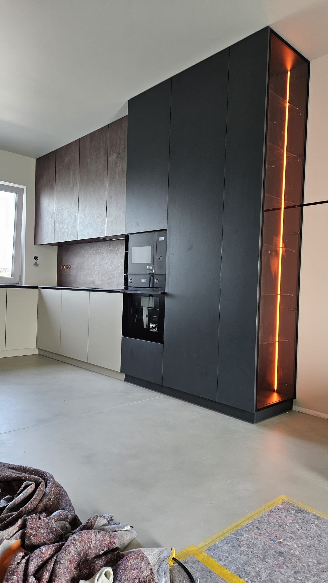 Nowoczesna zabudowa kuchenna z szafkami w kolorze ciemnego grafitu i beżu, z podświetlaną witryną i wbudowanymi sprzętami AGD, widoczne zabezpieczenia podłogi po montażu.