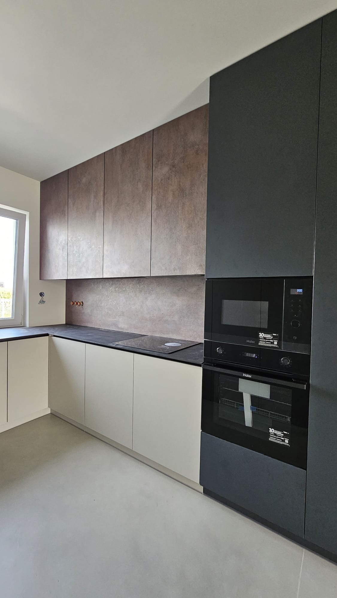 Nowoczesna kuchnia z górnymi szafkami w kolorze rdzy i dolnymi w odcieniach bieli, z czarnym blatem i zabudowanym piekarnikiem oraz mikrofalówką marki Haier.