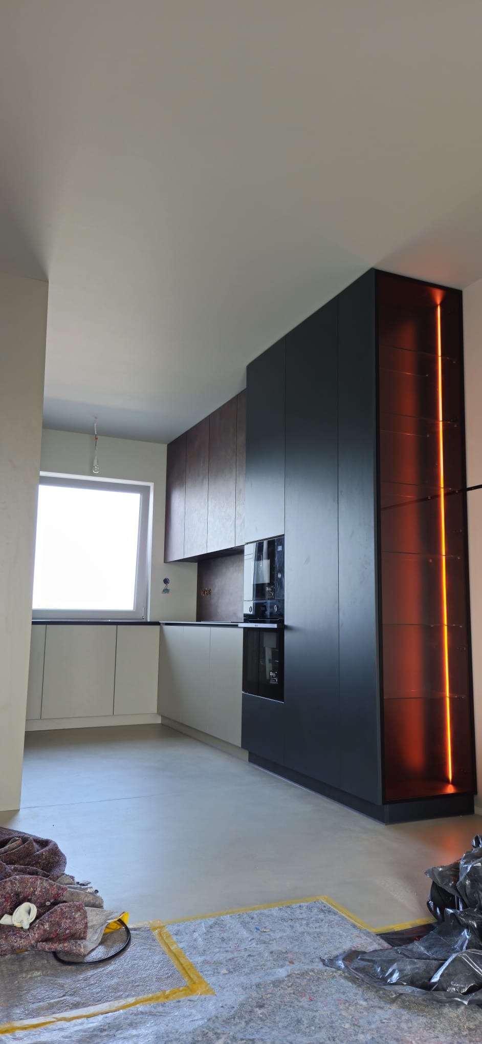 Nowoczesna kuchnia z szarymi i brązowymi szafkami, wbudowanym piekarnikiem i oświetloną pionową witryną, na podłodze widoczne materiały zabezpieczające podczas montażu.