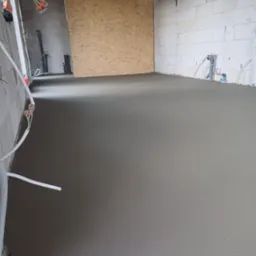 Świeżo wylana, gładka posadzka w surowym wnętrzu z białych bloczków, widoczne instalacje elektryczne i hydrauliczne w ścianach.