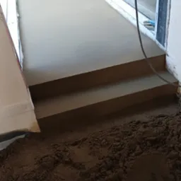 Świeżo wylana, gładka posadzka jastrychowa z dwoma stopniami, widoczny kabel zasilający i piasek w tle, przy oknie.