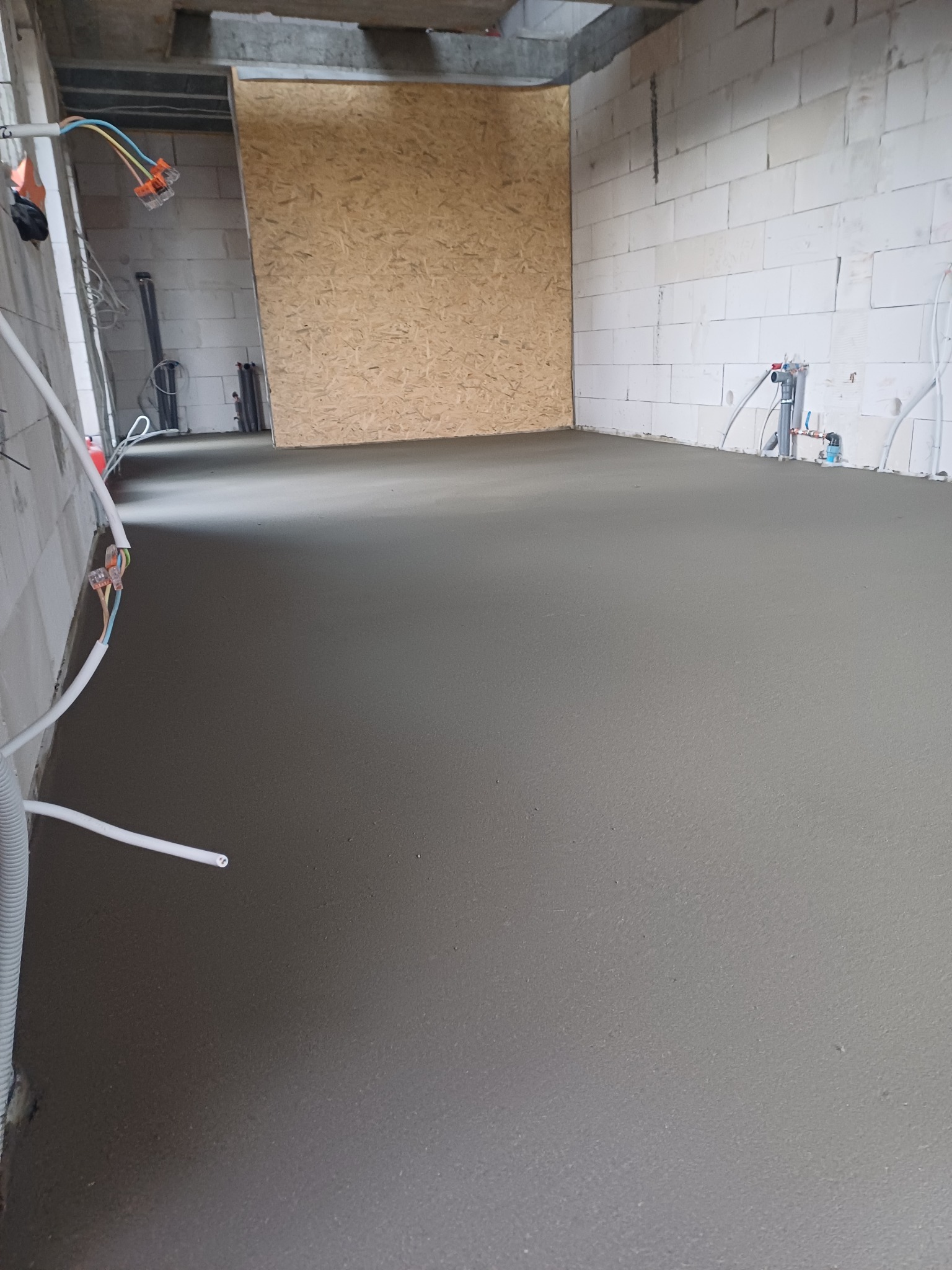 Świeżo wylana, gładka posadzka w surowym wnętrzu z białych bloczków, widoczne instalacje elektryczne i hydrauliczne w ścianach.