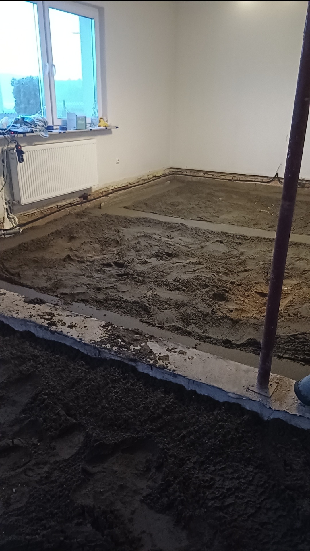 Przygotowanie podłoża pod wylewkę w pomieszczeniu z oknem, widoczne ślady stóp na mokrej powierzchni.