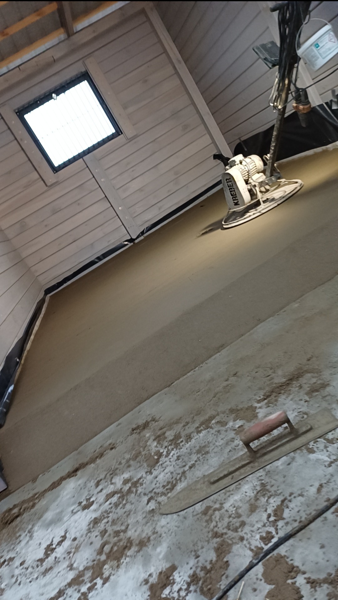 Wyrównywanie wylewki maszyną Kreber w pomieszczeniu z drewnianym stropem i oknem dachowym, widoczna packa na podłodze.