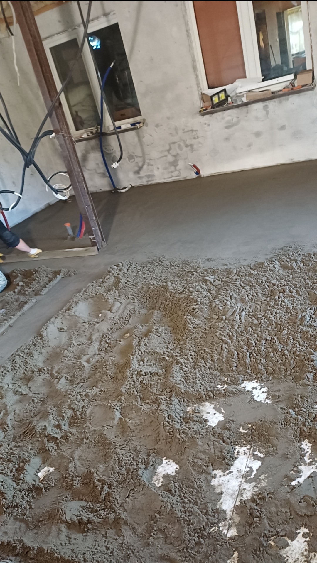 Wylewka podłogowa w trakcie wykonywania, widoczne częściowo rozprowadzone mokre podłoże i pozostała część w formie sypkiej mieszanki, ślady stóp na podłożu.