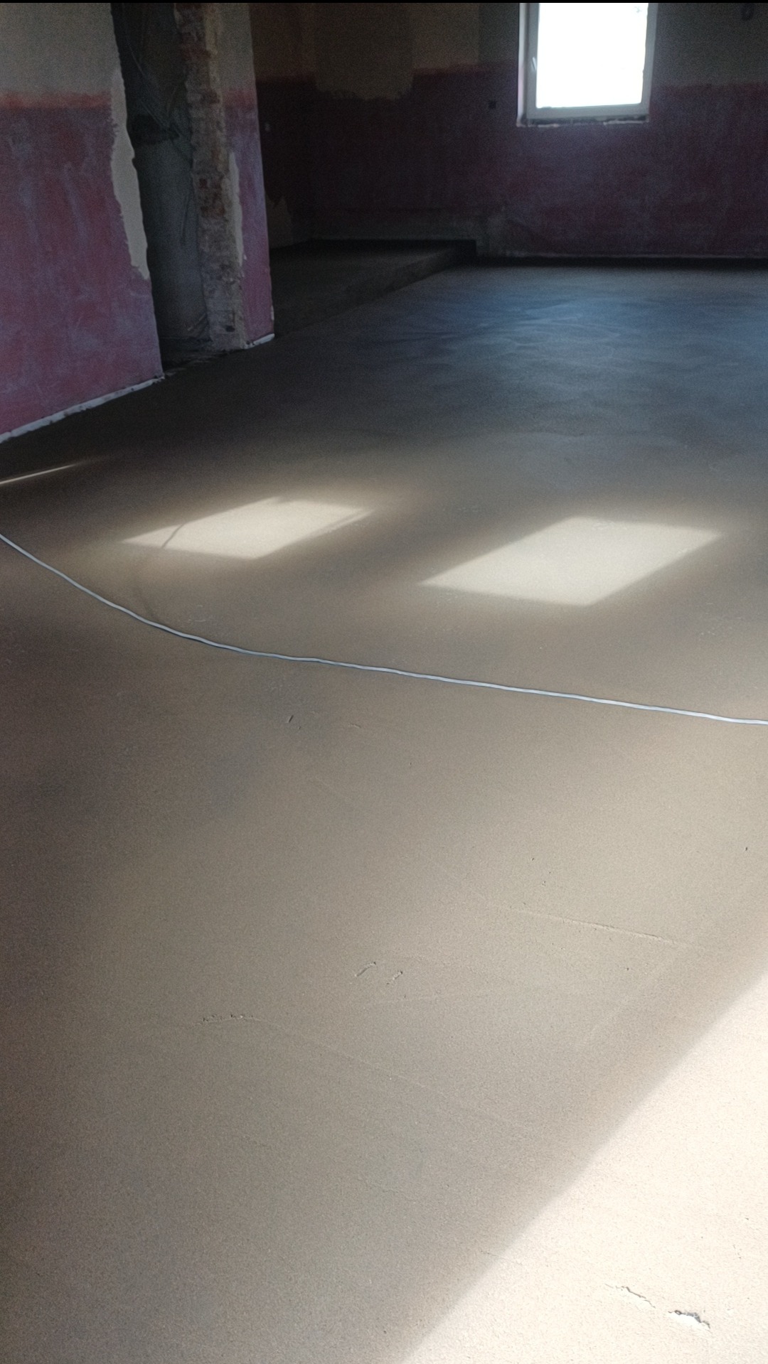 Świeżo wylana, gładka posadzka w surowym wnętrzu z widocznymi śladami po szpachli i smugami światła słonecznego wpadającego przez okno.