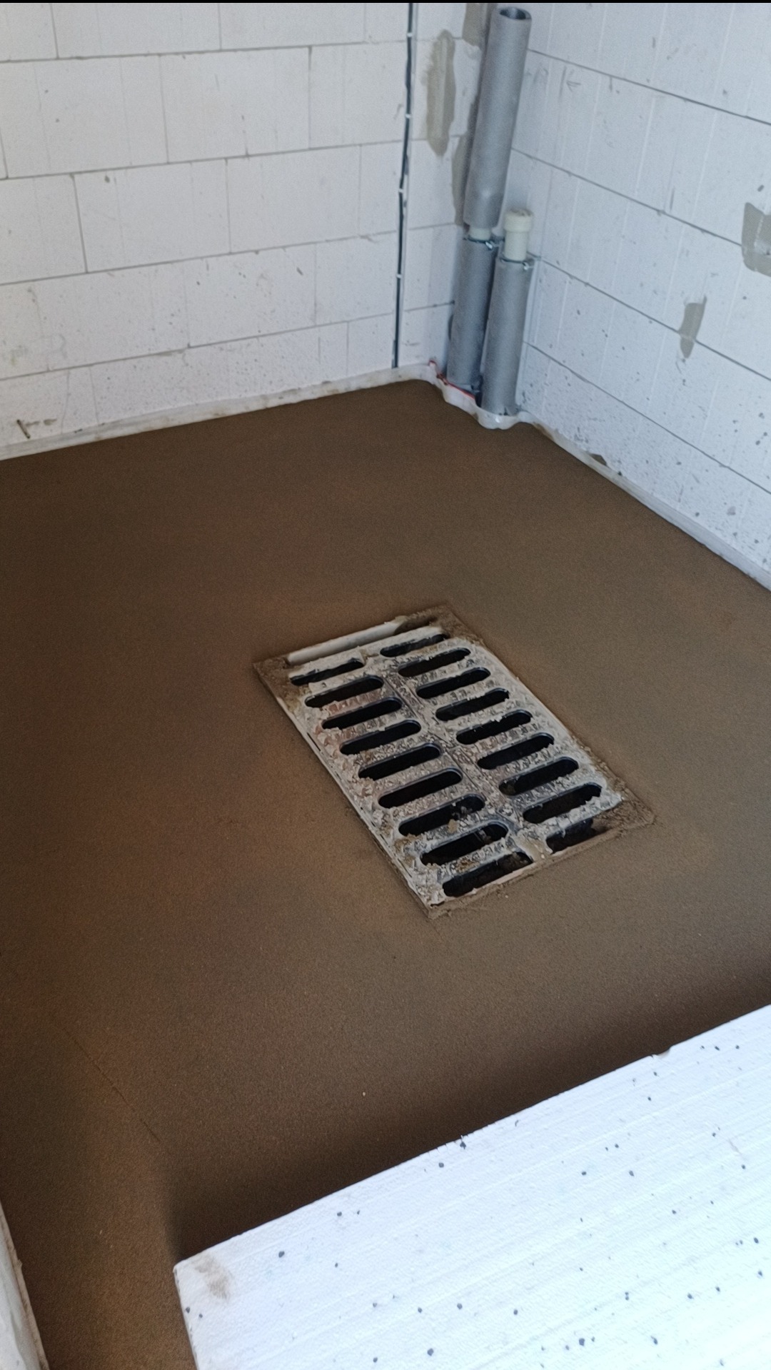 Świeżo wylana, brązowa posadzka z wbudowaną kratką ściekową w pomieszczeniu z białych bloczków i pionowymi rurami. Widoczne niedoskonałości i zabrudzenia na kratce.