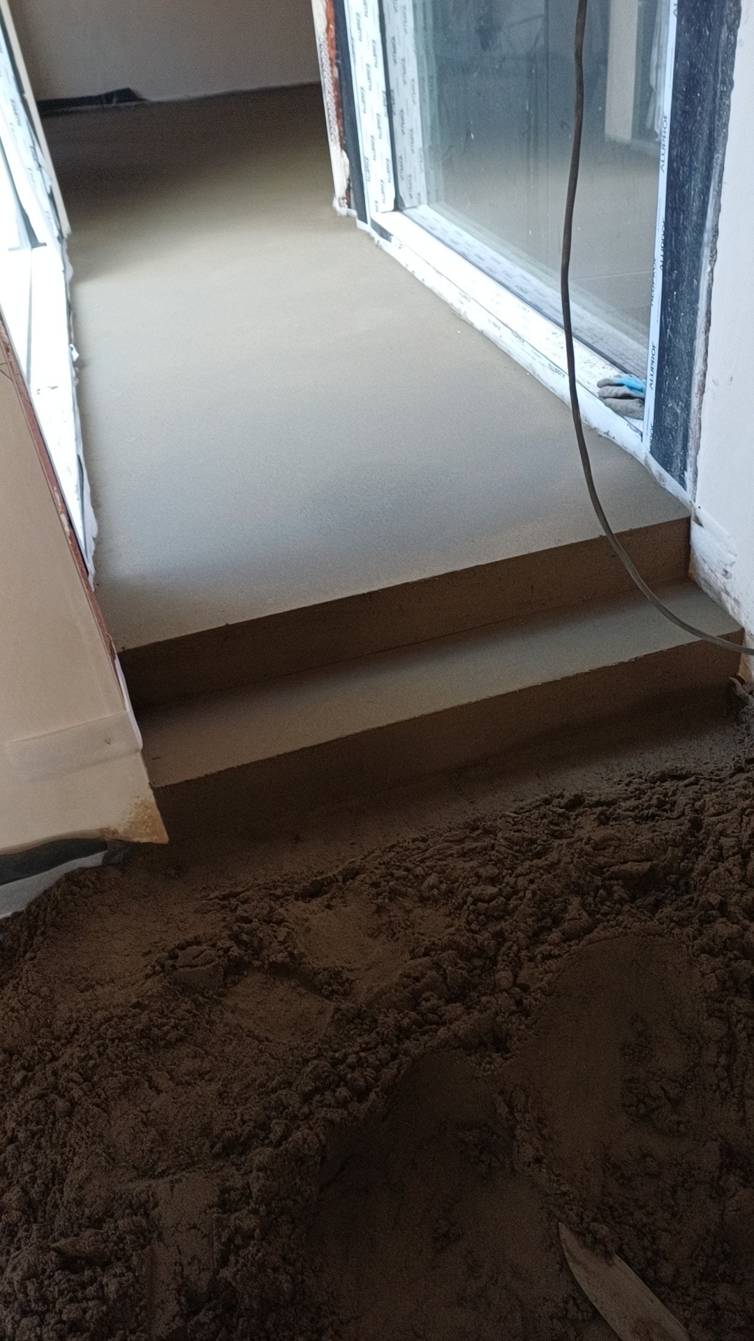 Świeżo wylana, gładka posadzka jastrychowa z dwoma stopniami, widoczny kabel zasilający i piasek w tle, przy oknie.
