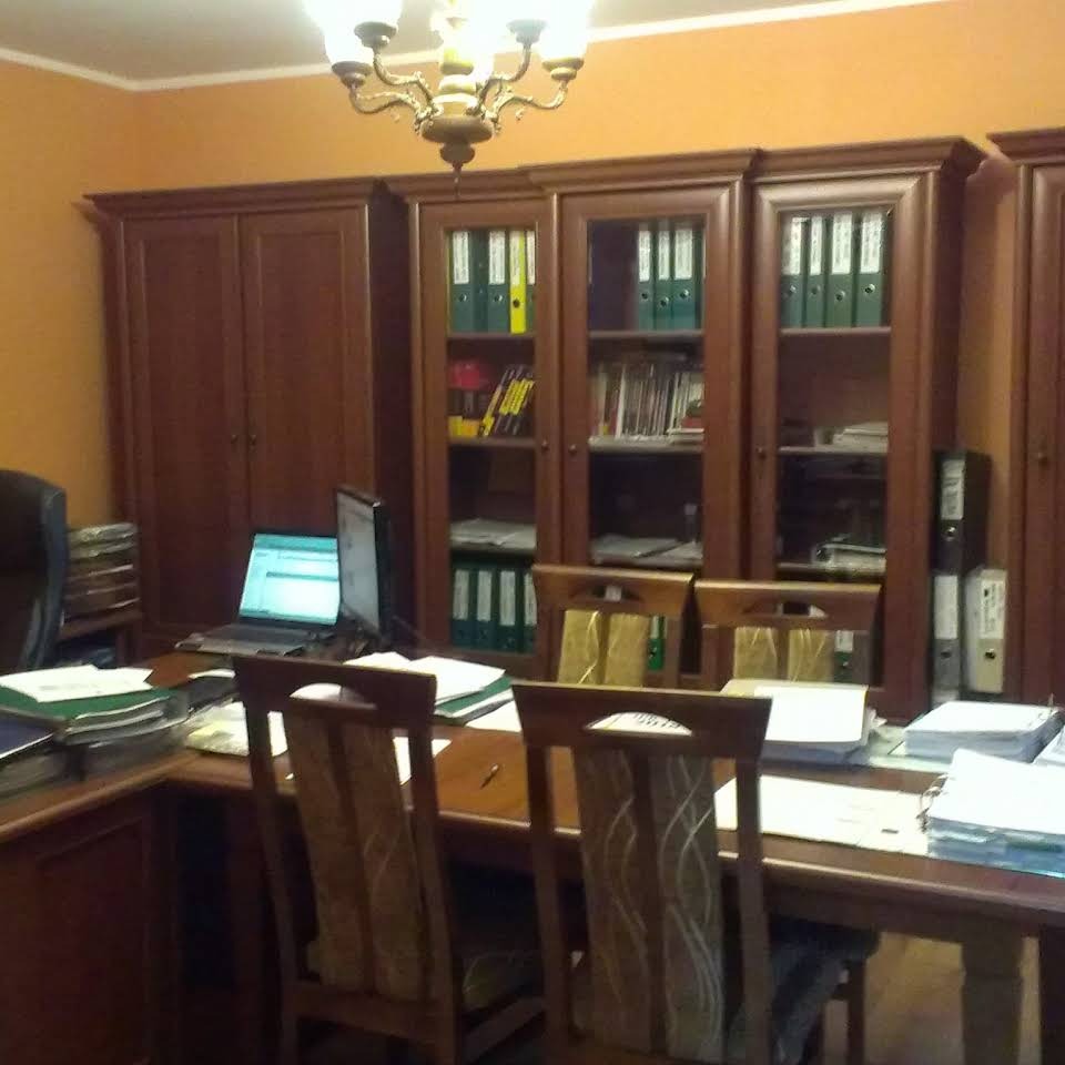 Biuro z drewnianymi meblami, regałami wypełnionymi segregatorami i książkami, biurkiem z laptopem i stosem dokumentów, oświetlone przez ozdobny żyrandol.