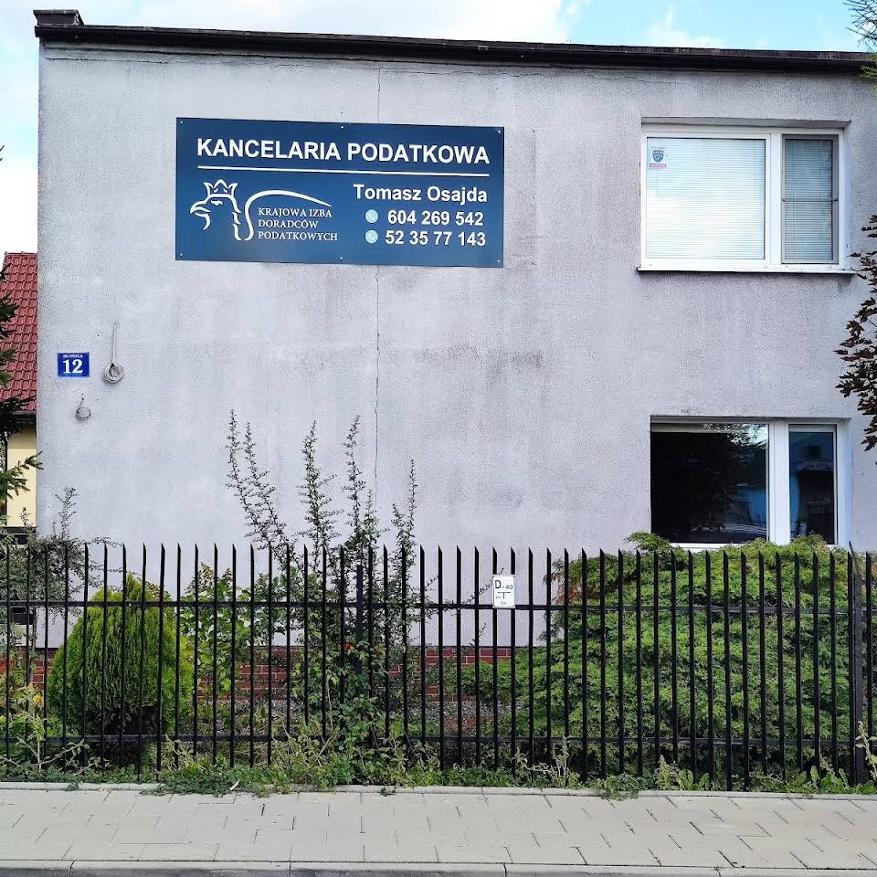 Szary budynek z tablicą informacyjną 'Kancelaria Podatkowa' z numerem telefonu, widok zza czarnego, metalowego ogrodzenia porośniętego zielenią.