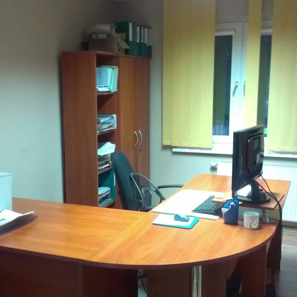 Biuro z biurkiem w kształcie litery U, komputerem, szafą z dokumentami i żółtymi żaluzjami w oknie.