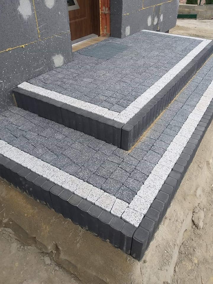 Schody wejściowe do domu wyłożone szarą kostką brukową z białym obramowaniem, widoczne niedokończone ocieplenie budynku.