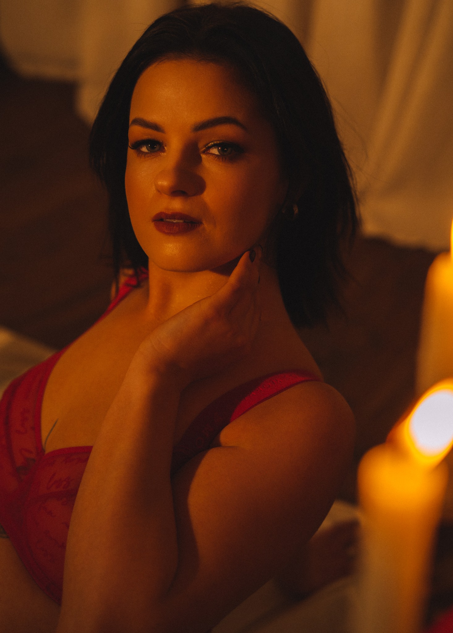 Portret kobiety w czerwonej bieliźnie, oświetlonej ciepłym światłem świec, z delikatnym makijażem i zamyślonym spojrzeniem, dłonią dotykającej szyi.