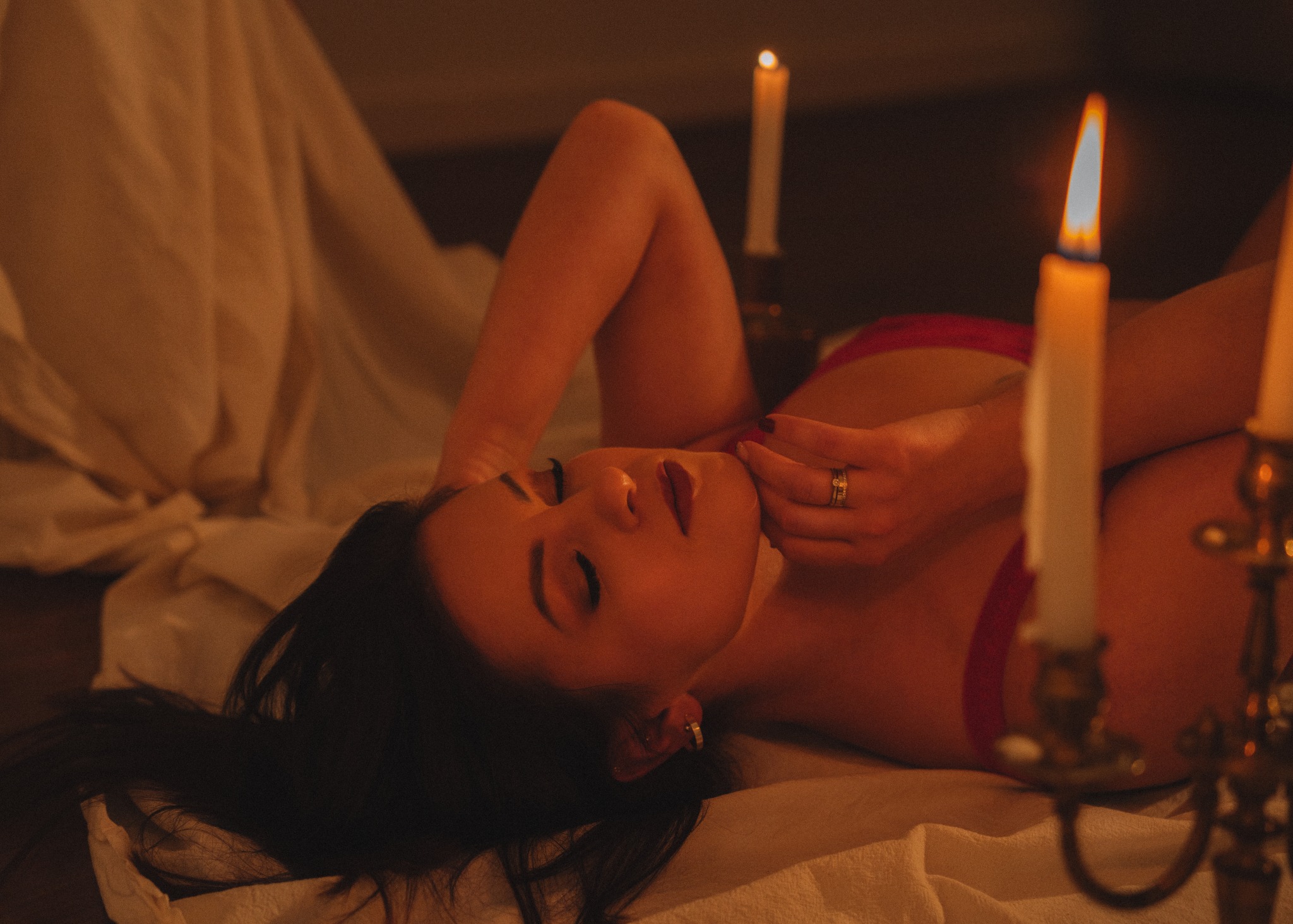Kobieta leży na białym materiale, oświetlona ciepłym światłem świec w ozdobnych świecznikach. Ujęcie z góry, skupione na twarzy i dłoniach.