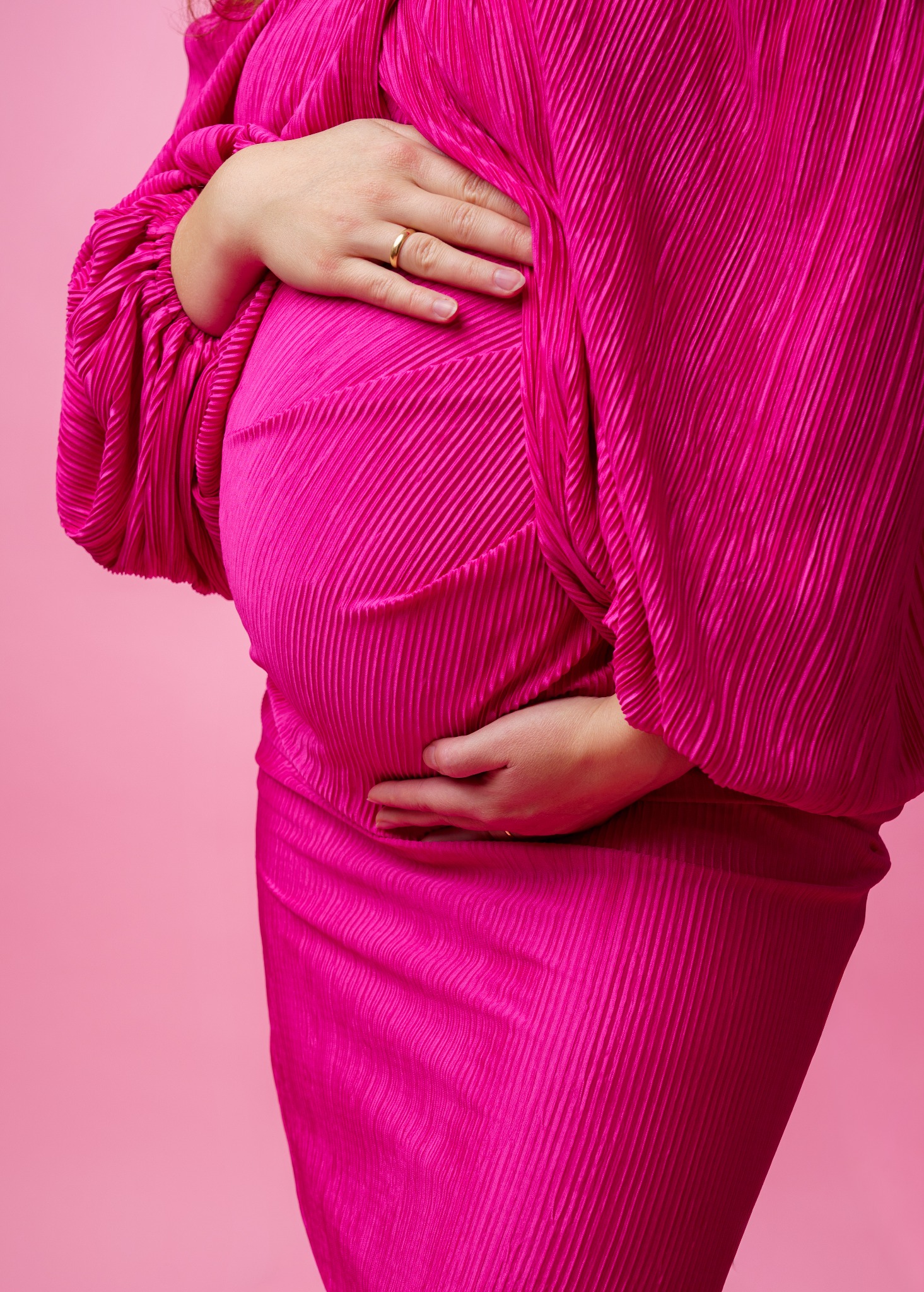 Zbliżenie na ciążowy brzuszek kobiety w plisowanej, fuksjowej sukience, dłonie spoczywają na brzuchu, na palcu widoczny złoty pierścionek, różowe tło.
