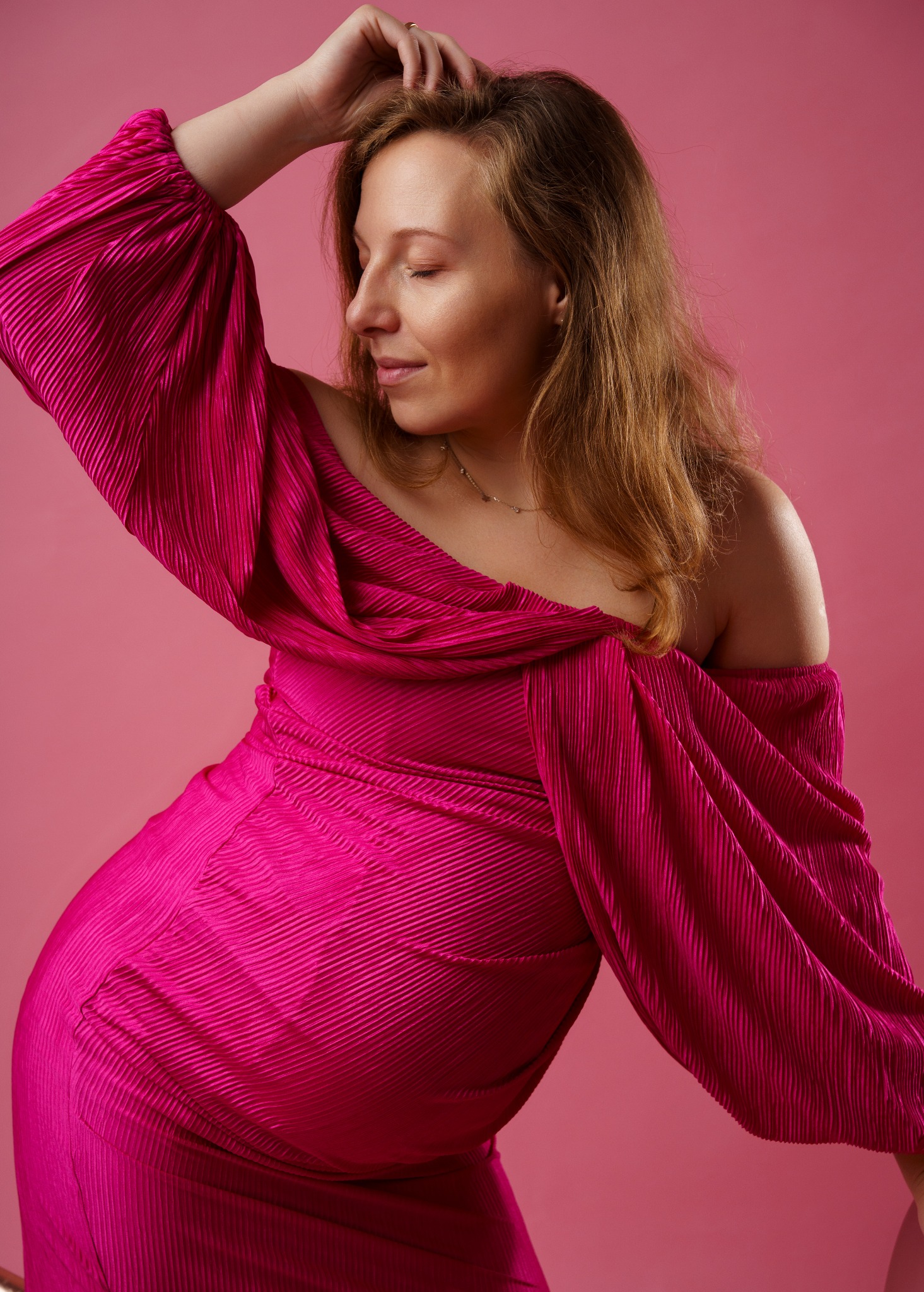 Portret kobiety w plisowanej sukni fuksjowej, z zamkniętymi oczami i dłonią we włosach, na jednolitym różowym tle. Ujęcie studyjne, miękkie światło.
