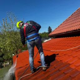 Pracownik w kasku i uprzęży myje czerwoną dachówkę z użyciem myjki ciśnieniowej. Widoczne strumienie wody i czyszczona powierzchnia dachu. Błękitne niebo w tle.