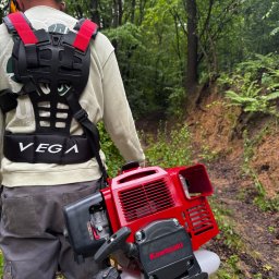 CleanTrans Bartosz Leśniowski - Mężczyzna z szelkami i kosą spalinową Kawasaki na tle zalesionego terenu. Widoczna ścieżka i naturalne otoczenie, sugerujące prace porządkowe w terenie.