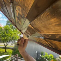 Malowanie drewnianej podbitki dachowej pędzlem, widok z dołu na tle zieleni. Prace renowacyjne z bliska, podkreślona faktura drewna i detale malowania.