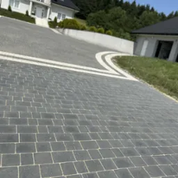 Ułożona, szara kostka brukowa na podjeździe do domu z białymi elementami dekoracyjnymi i zielenią w tle, w słoneczny dzień. Widok z perspektywy chodnika.