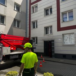 CleanTrans Bartosz Leśniowski - Pracownik w kasku i jaskrawej koszulce stoi przed budynkiem, obok podnośnika koszowego i pachołków, prawdopodobnie przygotowując się do mycia elewacji.