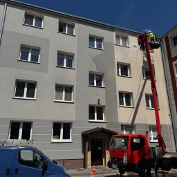 CleanTrans Bartosz Leśniowski - Pracownik w żółtym kombinezonie myje elewację budynku z podnośnika koszowego. Widoczny niebieski bus i czerwona ciężarówka. Na ziemi stoją pachołki.