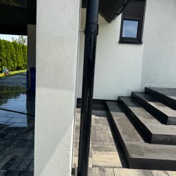 CleanTrans Bartosz Leśniowski - Świeżo umyta elewacja domu z czarną rynną i schodami z kostki brukowej. Widoczne mokre powierzchnie i sprzęt do mycia ciśnieniowego w tle.
