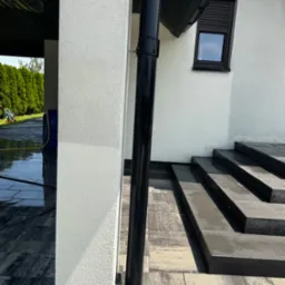 Świeżo umyta elewacja domu z czarną rynną i schodami z kostki brukowej. Widoczne mokre powierzchnie i sprzęt do mycia ciśnieniowego w tle.